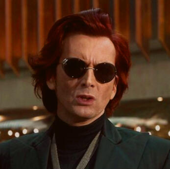 A headshot of Crowley from Good Omens. He's in a black turtleneck with a slim, gray jacket. He has red medium hair that goes down to about an inch over his shoulders.