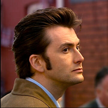 A headshot of The Doctor from Doctor Who. He's looking to the right with a neutral face. He has short, dark brown hair that swerves back. He wears a light blue collar shirt with a light brown jacket overtop.